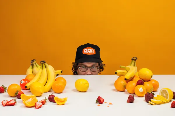 Man in a Mailchimp cap peeking over a table of fruit on orange