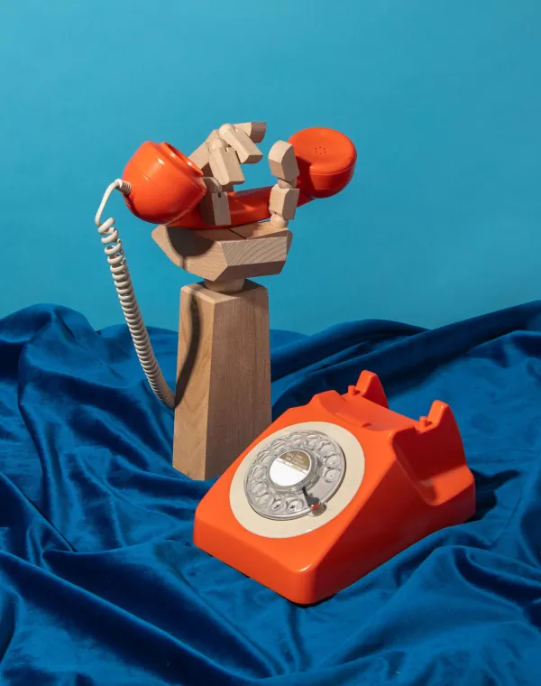 A carved wooden hand lifting an orange rotary phone receiver over blue fabric